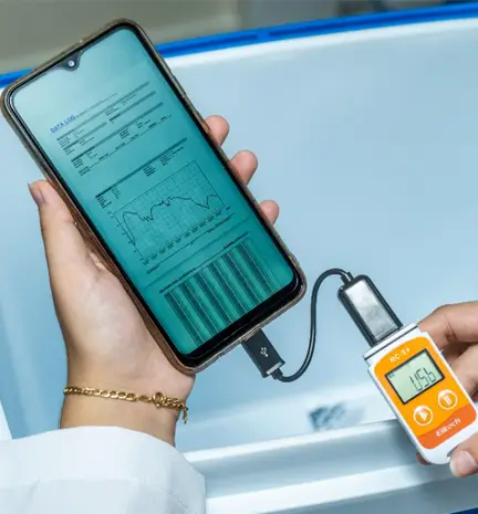 Uma mão com uma pulseira dourada segurando o celular que está conectado com um data logger de temperatura, mostrando dados e em cima de um suporte branco