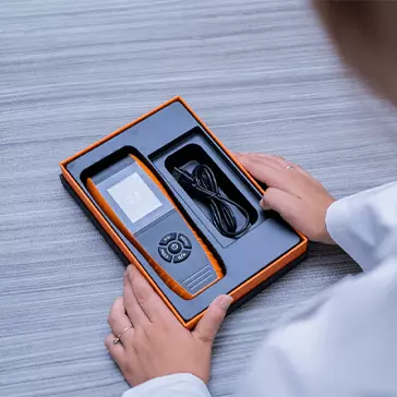 Detector de qualidade de ar dentro de uma caixa com seu acessório em cima de uma mesa. Uma pessoa está segurando a caixa com as duas mãos